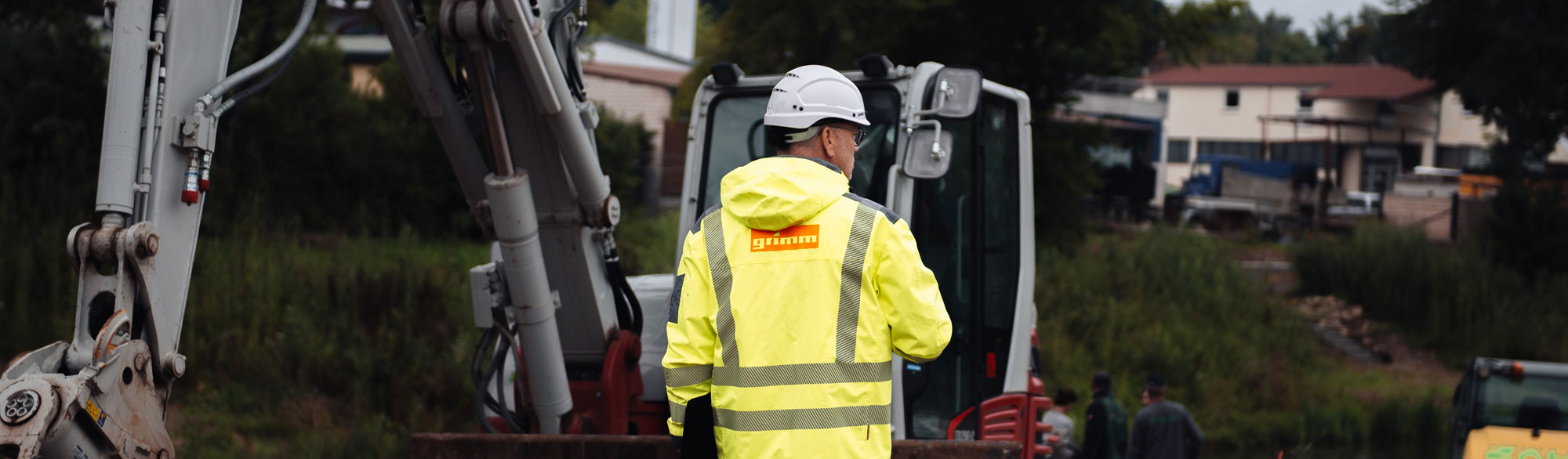 Ein Mann steht mit Arbeitsjacke und Helm auf einer Baustelle und überwacht die Arbeiten.